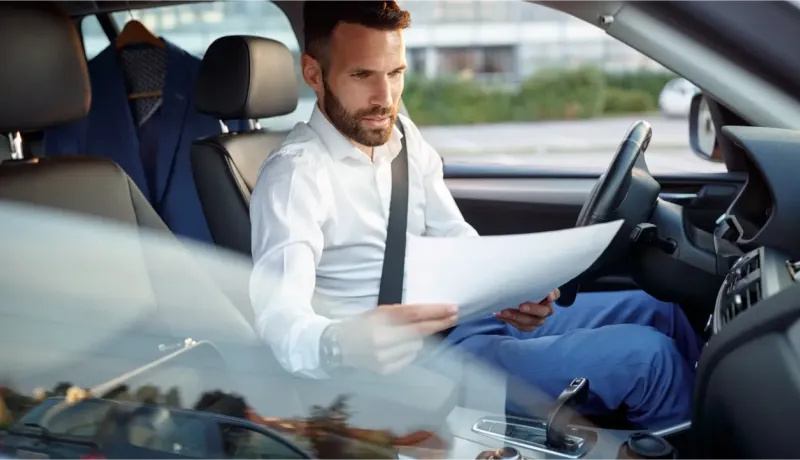 Un homme barbu en chemise lit des documents au volant d'une voiture, un costume à l'arrière.