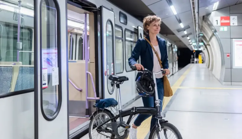 Une femme souriante sort d'un train dans une station de métro, tenant un vélo pliant.