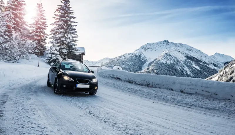 Une voiture sombre roule sur une route de montagne enneigée, sous un ciel ensoleillé.