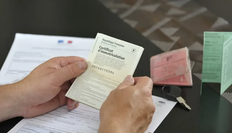 Mains tenant un certificat d'immatriculation français, avec d'autres documents et une clé.