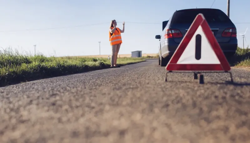 Un triangle de signalisation d'urgence sur la route devant une voiture en panne et une femme.