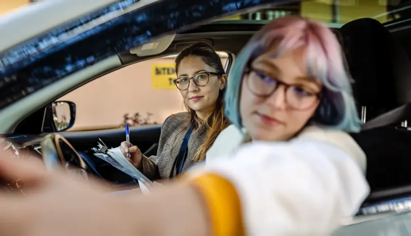 Deux femmes dans une voiture, une conductrice et l'autre tenant un presse-papiers, potentiellement un examen de conduite.