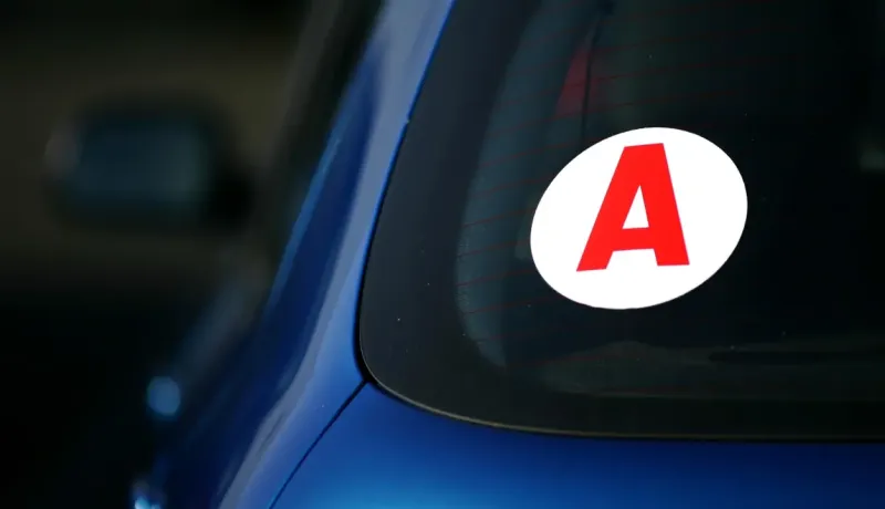 Gros plan d'un autocollant blanc avec un grand "A" rouge, signalant un jeune conducteur à l'arrière d'une voiture bleue.