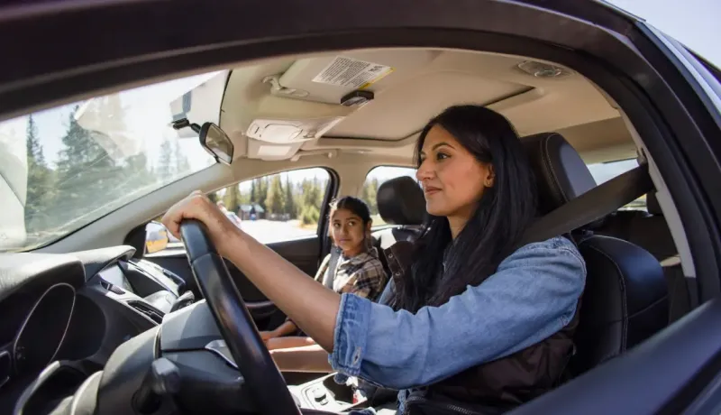 Une femme souriante conduit une voiture, sa fille assise sur le siège passager arrière, regardant au loin.