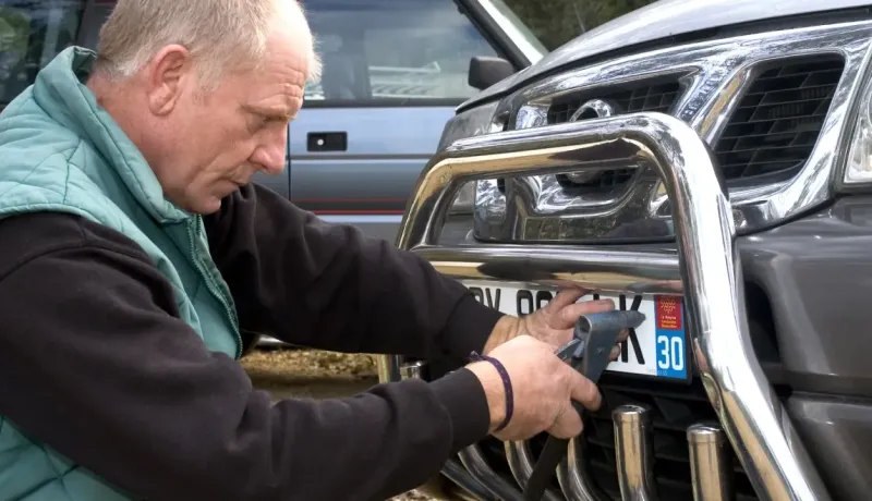 Un homme en veste verte fixe une plaque d'immatriculation sur le pare-buffle avant d'une voiture.