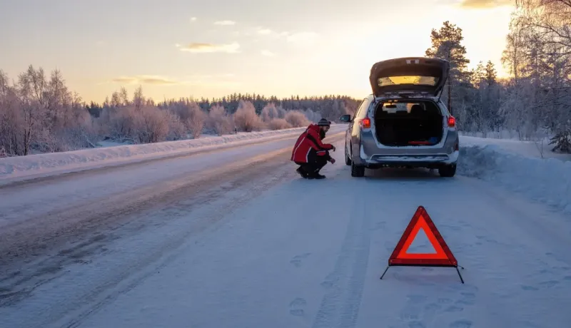 Une personne avec un gilet orange s'accroupit derrière une voiture en panne sur une route enneigée avec un triangle de signalisation.