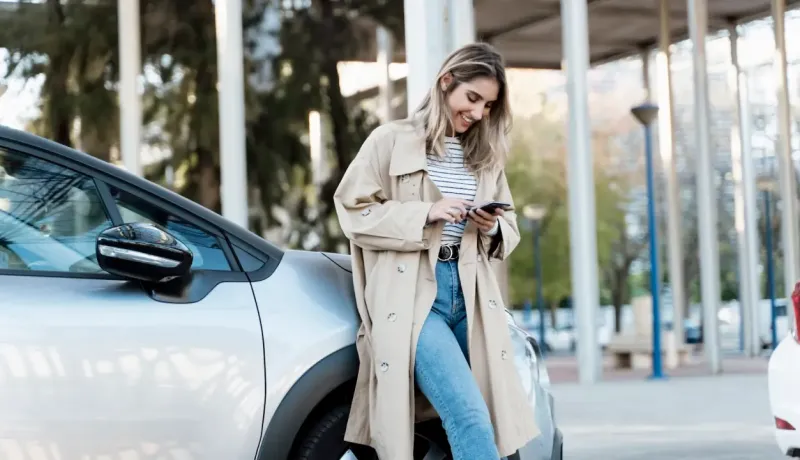 Une femme adossée à sa voiture consulte son téléphone.