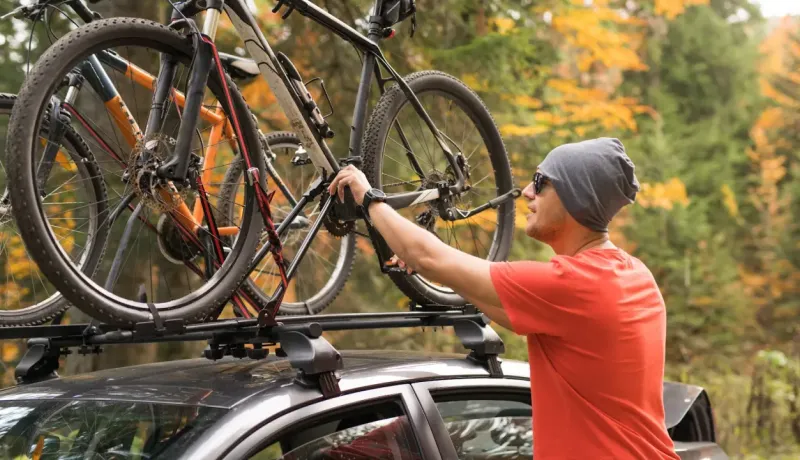 Un homme en t-shirt orange, bonnet et lunettes de soleil, attache un vélo sur le porte-vélo de sa voiture.