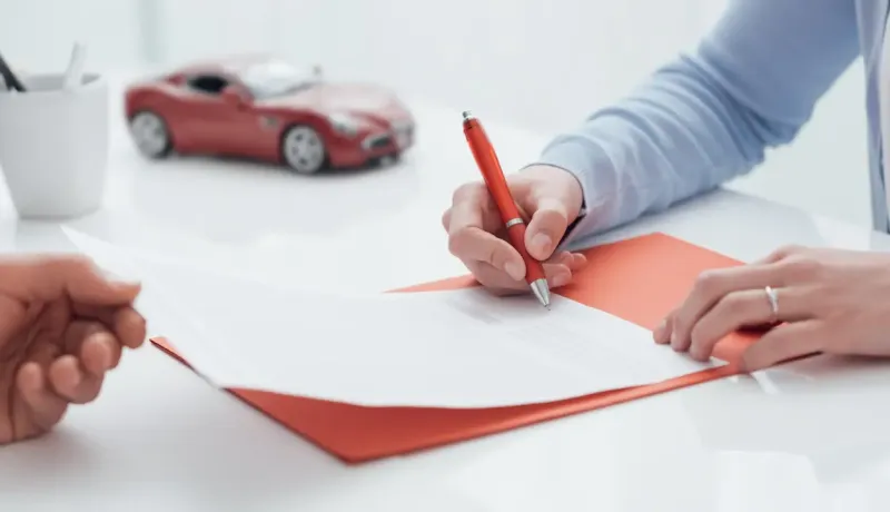 Deux personnes signent des documents d'assurance automobile avec un stylo rouge, une petite voiture rouge est visible en arrière-plan.