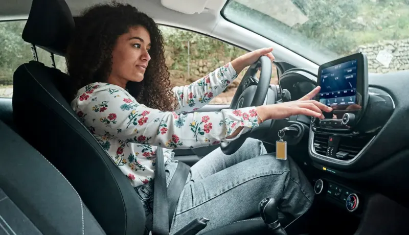 Jeune femme aux cheveux bouclés, assise au volant, interagissant avec l'écran tactile du système de bord.