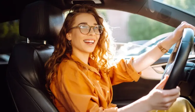 Jeune femme souriante aux cheveux bouclés et lunettes, conduisant une voiture.