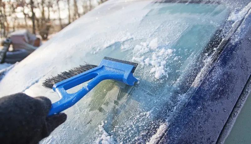 Une main gantée gratte le givre d'un pare-brise de voiture avec un grattoir à neige bleu.