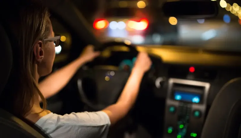 Une femme portant des lunettes conduit une voiture la nuit, les lumières de la ville floues en arrière-plan.