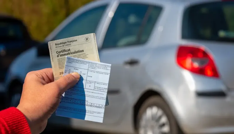 Une main tenant un certificat d'immatriculation et d'autres documents, devant une voiture en arrière-plan.