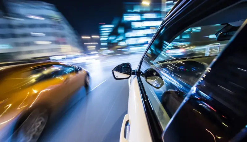 Vue latérale d'une voiture sur une route, avec une autre voiture rapide et les lumières floues de la ville la nuit.