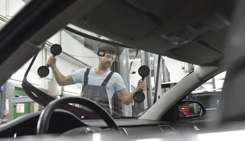 Un ouvrier installe un nouveau pare-brise sur une voiture dans un garage, vu de l'intérieur de la voiture.