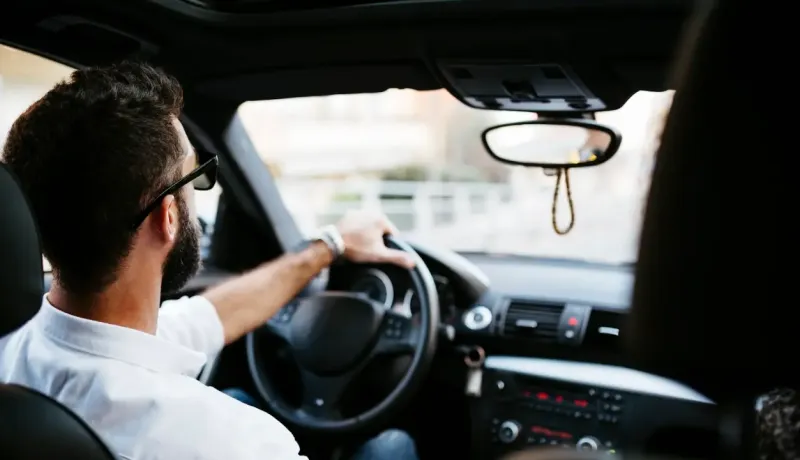 Un homme barbu portant des lunettes de soleil conduit une voiture, vu de derrière son épaule.