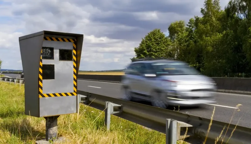 Un radar de vitesse gris et jaune sur le bord d'une autoroute, avec une voiture floue en mouvement.