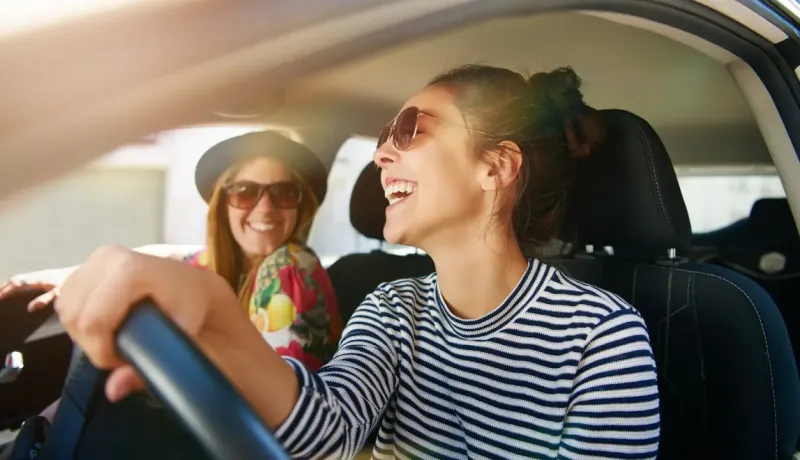 Deux jeunes femmes souriantes et heureuses dans une voiture, la conductrice rit aux éclats.