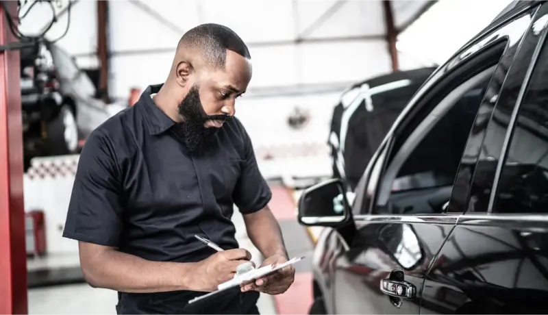 Un mécanicien barbu examine des documents à côté d'une voiture noire dans un garage.
