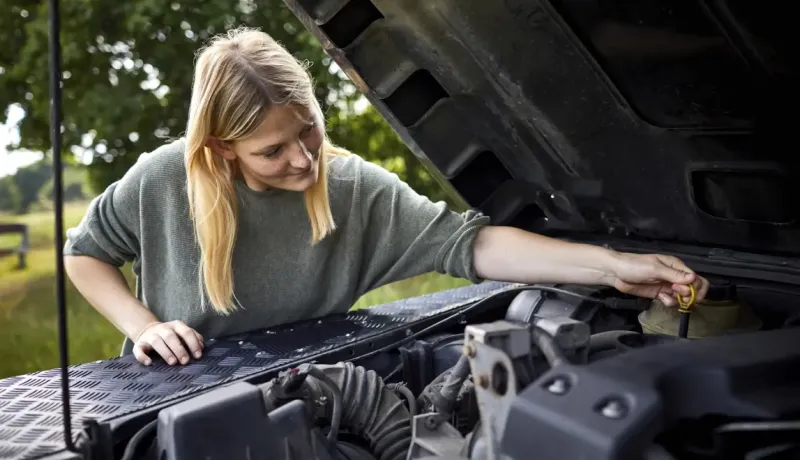 Une jeune femme aux cheveux blonds vérifie le niveau d'huile du moteur d'une voiture, le capot ouvert.