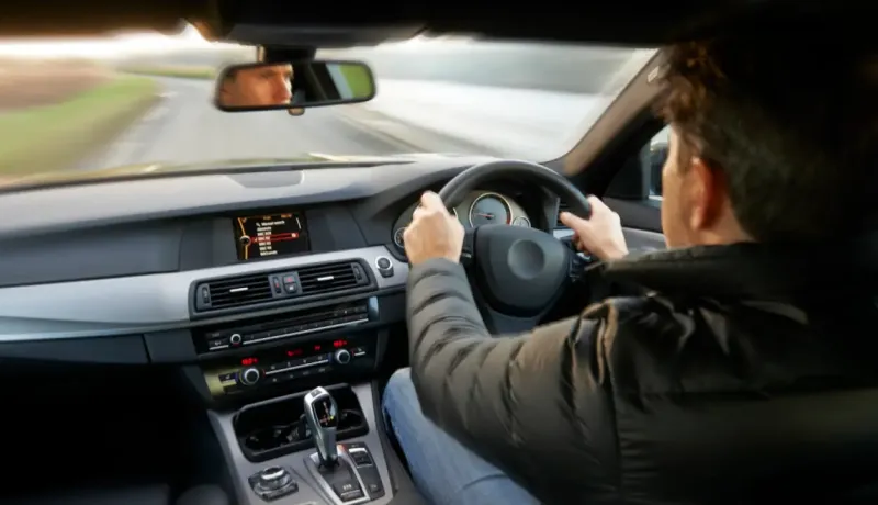 Vue depuis l'intérieur d'une voiture, un homme conduit sur une route de campagne, son reflet dans le rétroviseur.