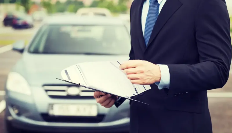 Un homme en costume tient un dossier de documents devant une voiture garée, évoquant l'assurance.