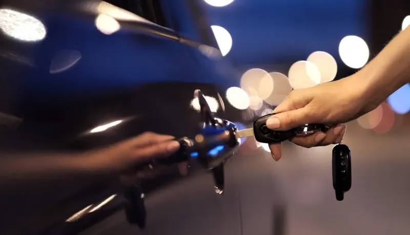 Une main ouvre la porte d'une voiture noire avec une clé, les lumières de la ville floues en arrière-plan.