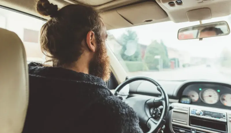 Un homme barbu avec un chignon conduit une voiture, vu de dos, regardant la route devant lui.