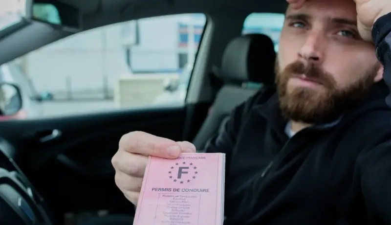 Un homme barbu au regard perplexe tient un permis de conduire français rose vif dans une voiture.