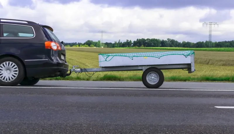 Une voiture noire tracte une petite remorque avec un filet vert sur une route, un champ en arrière-plan.