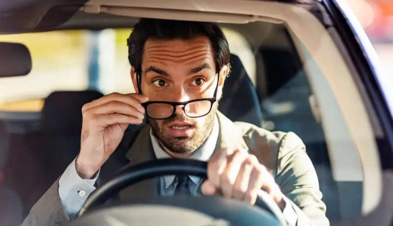 Un homme en costume, l'air surpris, retire ses lunettes en conduisant.