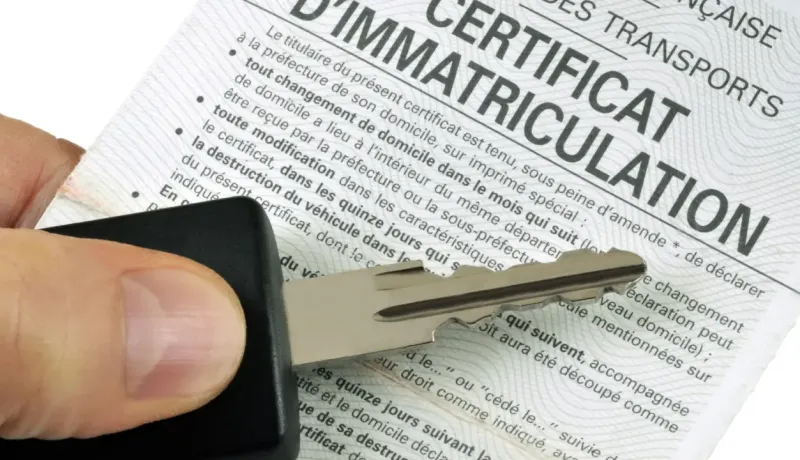 Une main tient une clé de voiture au-dessus d'un certificat d'immatriculation de voiture français.