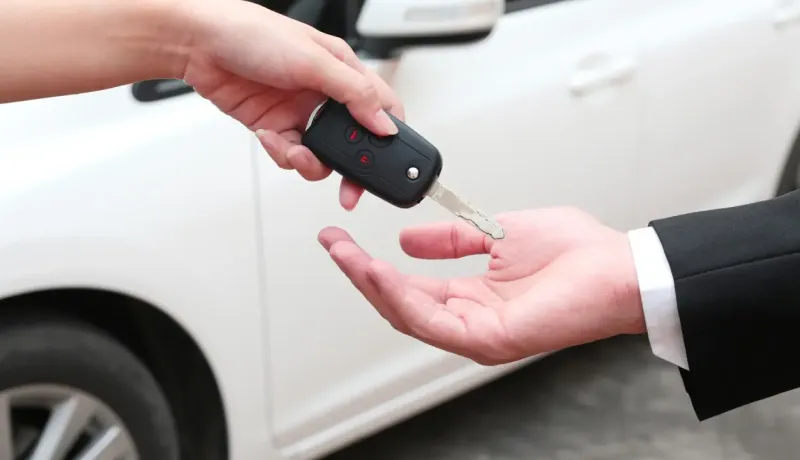 Deux mains échangent une clé de voiture devant un véhicule blanc, symbolisant le partage d'une voiture entre particuliers.