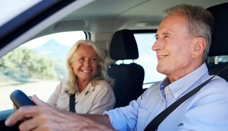 Un couple âgé souriant est assis dans une voiture, l'homme conduit et la femme le regarde.