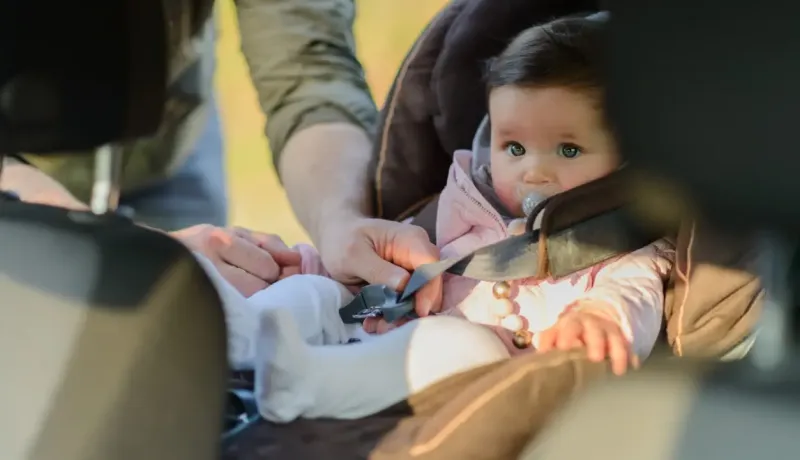 Un adulte ajuste les sangles d'un bébé assis dans un siège auto, le bébé regarde vers l'avant.