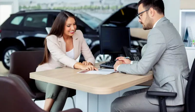 Une femme et un homme discutent de documents à un bureau dans ce qui semble être une concession automobile.