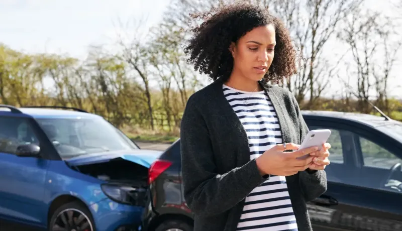 Une femme avec des cheveux bouclés regarde son téléphone, avec deux voitures accidentées en arrière-plan.