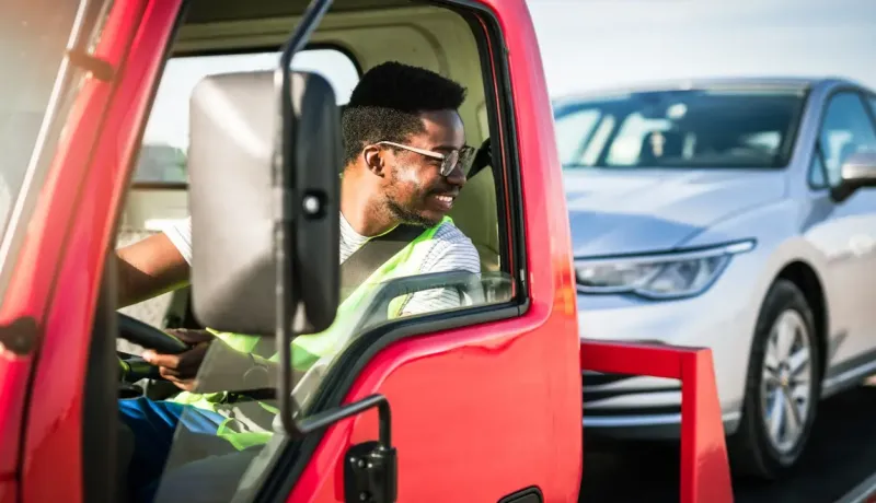 Un conducteur souriant dans un dépanneuse rouge, avec une voiture argentée à l'arrière.