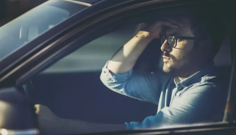 Un homme l'air préoccupé ou triste, la main sur le front, est assis au volant d'une voiture.