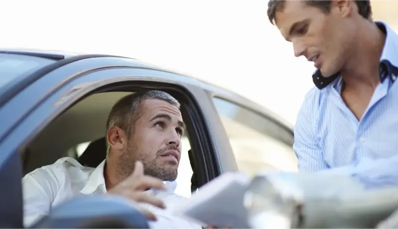 Deux hommes discutent à côté d'une voiture, l'un à l'intérieur, l'autre à l'extérieur.