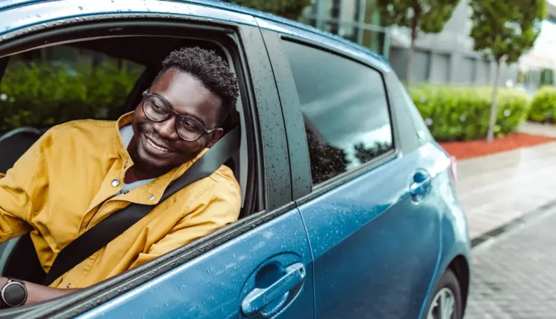 Un homme souriant dans une voiture, le regard tourné vers l'avant, avec des gouttes de pluie sur la carrosserie.