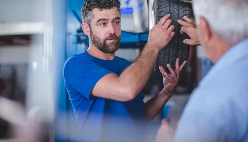 Un mécanicien barbu en blouse bleue explique un pneu à un client dans un garage.