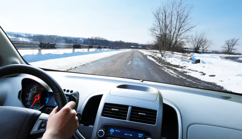 Vue du siège conducteur d'une voiture roulant sur une route enneigée avec des arbres dénudés.