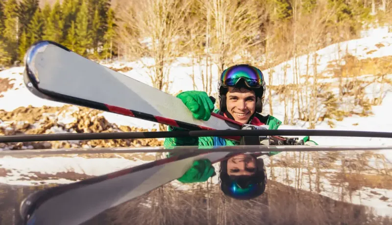 Un skieur souriant attache des skis sur le toit d'une voiture, avec un paysage hivernal en arrière-plan.