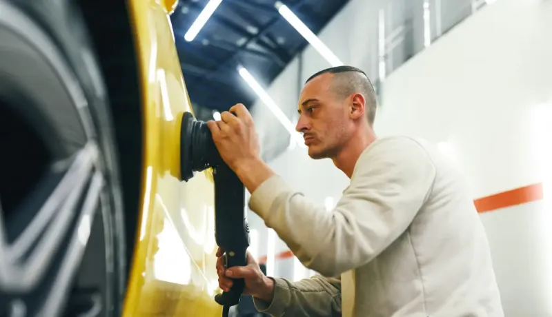 Un homme travaille méticuleusement sur la carrosserie d'une voiture jaune avec une polisseuse.