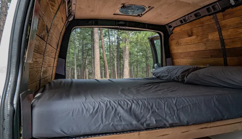L'intérieur d'une camionnette aménagée en camping-car avec un lit et vue sur une forêt.