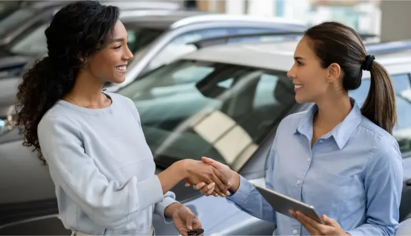Deux femmes se serrent la main souriantes, une tenant des clés de voiture, dans une concession.