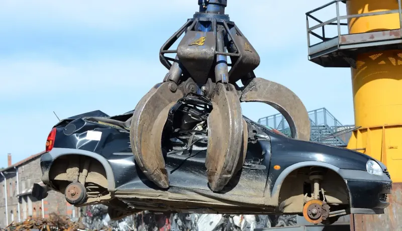 Une grue avec une pince géante soulève une voiture noire fortement endommagée dans une casse.