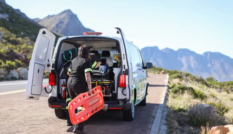 Deux ambulanciers se préparent à sortir leur équipement, dont une planche dorsale orange, d'une ambulance garée au bord d'une route de montagne.